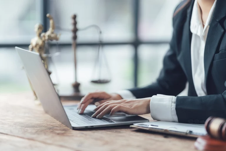 Eine Anwältin sitzt an ihrem Laptop. Im Hintergrund sieht man eine alte Waage und im Vordergrund liegt ein Richterhammer.