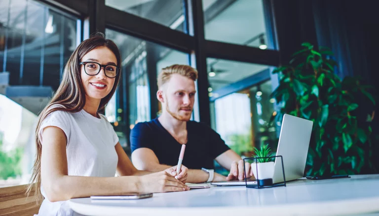 Eine Frau sitzt lächelnd an einem Tisch. Sie hält einen Stift in der Hand und macht sich Notizen, während sie freundlich in die Kamera schaut. Der Mann arbeitet im Hintergrund an seiner Online-Präsenz am Laptop.