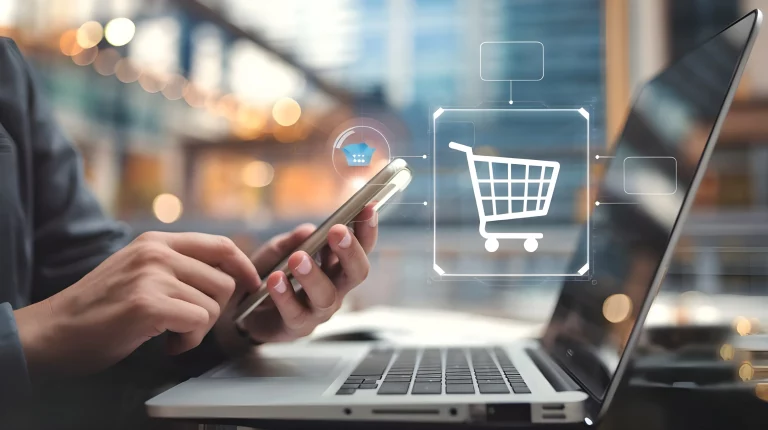 Eine Person sitzt mit ihrem Handy an einem Laptop und kauf online etwas. Im Vordergrund sieht man ein Symbol eines Einkaufswagens um den Kauf darzustellen.