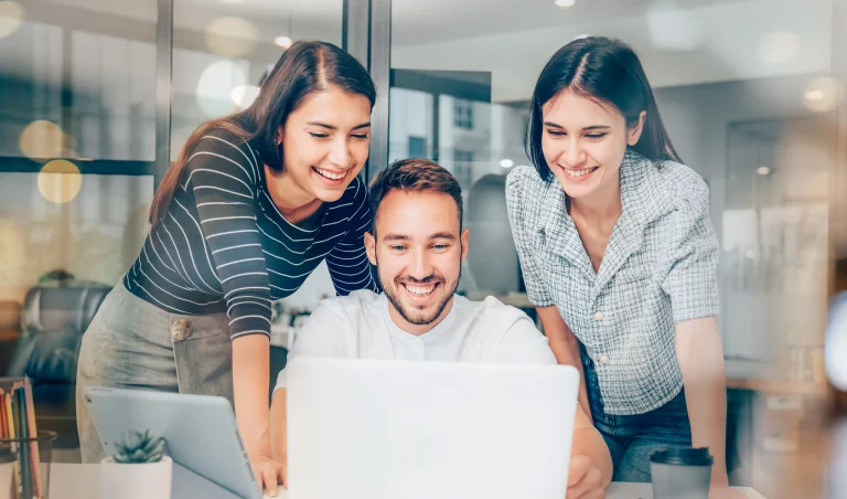 Ein Mann sitzt lachend vor seinem Laptop. Seine 2 Kolleginnen stehen um ihn herum und lachen auch, während Sie auf den Laptop schauen. Sie freuen sich über ihre gute Sortierung der Dokumente in ihrem DMS - Dokumenten-Management-System.